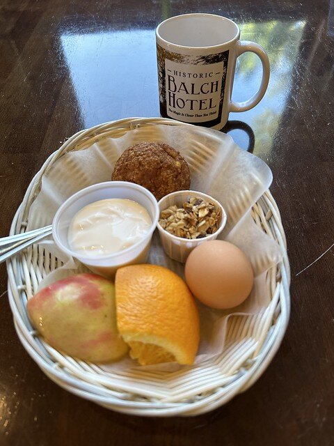 Balch Hotel breakfast basket contains muffin, vanilla yogurt and small paper container with granola, brown hard boiled egg, orange section with peel, fuji apple quarter with skin in straw basket with wax paper sheet.