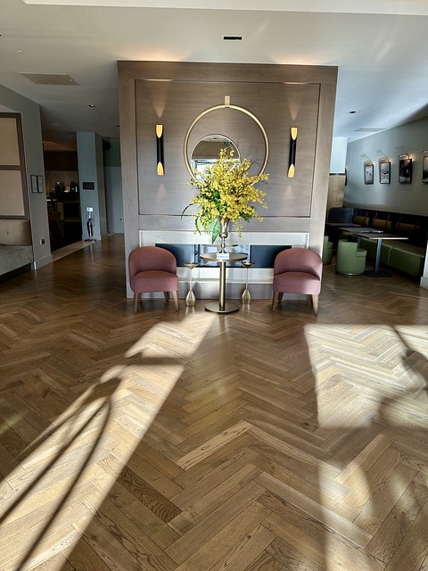 Light-filled hotel lobby with yellow floral display. Two chairs are next to the wall that separates the lobby from a gathering area. The floor is inlayed wood. 
