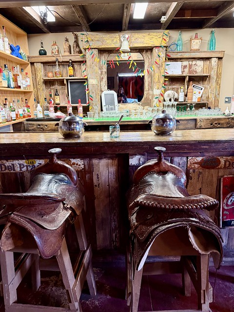 Three Western saddles serve as bar stools, perched on wood bases in the historic cantina. Two silver-plated, circular ice buckets sit on the wood bar with a metal ice scoop in a small glass container between the buckets. Numerous bottles of spirits and cocktail glasses line the shelves in the ranch bar.