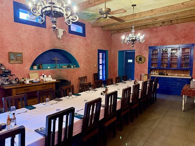 Red painted walls and bright blue painted door and 2 rectangular windows frame the room. A blue painted, built-in buffet holds glasses in a cupboard with 3 drawers below the countertop. A Mexican sombrero hat hangs on the wall to the left of the cupboard and right of the blue painted kitchen door. A cow skull is mounted on the wall, below the rectangular 2 windows. A built-in alcove, above the wood burning fireplace, holds a cross, 2 small ceramic water jugs and small artifacts from the 1800's. A long wood table, with white linen table cloth, holds 18 wood chairs, 9 on either side of the long rectangular table. The table is set with blue napkins, silverware and water glasses for dinner service.