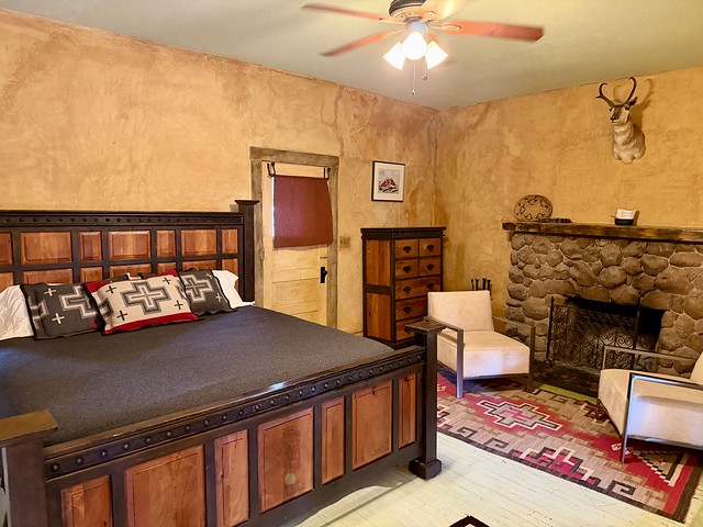 Partially shown is the king bed with dark wood head and base board. A black comforter covers the bed with 3 small rectangular pillows with western-themed decorative throw pillows on the bed. Two white cloth and wrought-iron chairs are positioned in front of the wood-burning fireplace. A red, white, black and beige Navajo area rug is under the 2 heavy chairs by the river rock fireplace. A taxidermy antelope head is mounted above the fireplace. Beige wall color on all walls. Back door is left of heavy wood dresser on the back wall in the bedroom. 