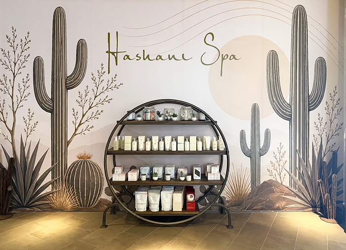 a circular shelf of products sits in front of a desert-themed mural with cactus. Hashani Spa logo is on wall above shelving.