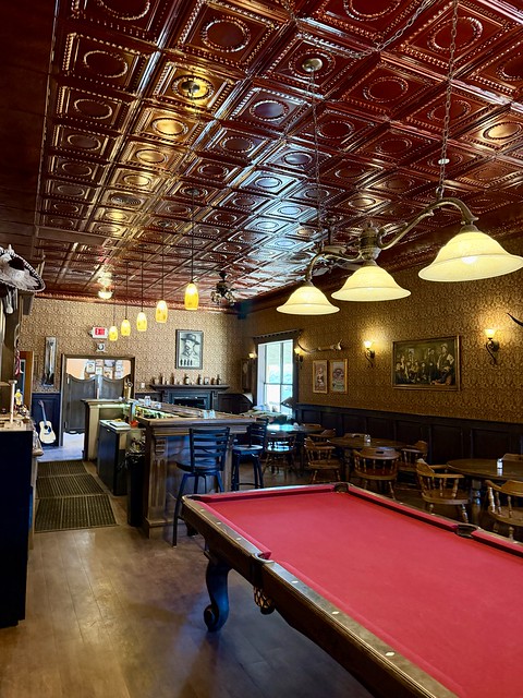 Hotel bar with faux brass ceiling squares, billiards table with red felt tablecloth. Three lamp brass fixture hangs over pool table. Beyond table is the wood bar with counter and bar stools. Across from the bar are 3 circular tables with 4 wood chairs around each table. A pencil sketch of Wyatt Earp is hanging in a frame above the metal fireplace, partially shown.