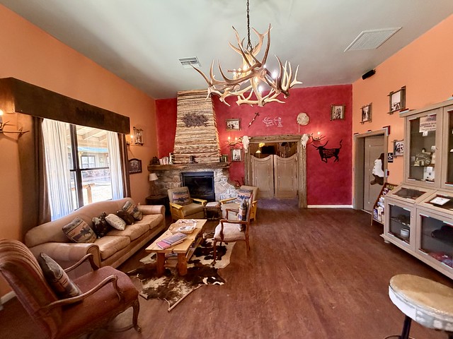 Ranch lobby has red painted back wall with river rock stone, wood burning fireplace. Right of fireplace are swinging wood doors that open to the saloon. Cow skulls frame either side of swinging doors. Wood floor has a brown and white cow hide on the floor with rectangular wood coffee table on top of cowhide rug. A beige leather sofa has 5 decorative, western-themed pillows on the couch. In front of the fireplace are 2 leather chairs with wood frames. Each chair has a western, decorative pillow. A mauve colored cloth chair, with wood arm handles is left of sofa. It, too, has a western decorative pillow. Across from the sofa is a glass and beige wood display case. A deer antler chandelier is hanging from the ceiling of the hotel lobby.